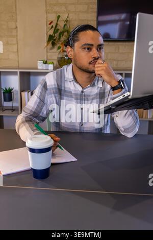 Erwachsener Mann, der am Schreibtisch im Büro sitzt, grünen Stift über das ausgekleidete Notebook hält und am Laptop arbeitet Stockfoto