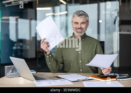Reife Führungskräfte oder Finanzexperten lächeln und halten Geschäftsdokumente in der Hand, während sie an einem modernen Schreibtisch mit Laptop und Papierkram arbeiten, und zeigen Erfolg und Positivität Stockfoto