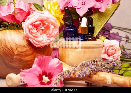 Flaschen mit ätherischem Öl in einem Holzmörtel umgeben von frischen Blumen und Kräutern, die natürliche Wellness und Aromatherapie repräsentieren. Stockfoto