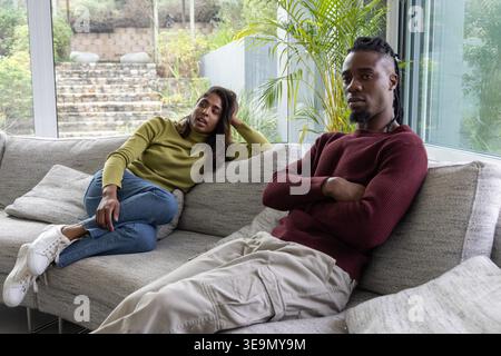 Verschiedene Paare sitzen angespannt auf einem hellgrauen Sofa mit neutralen Kissen und raumhohen Fenstern. Minimalistisch, modern, Tageslicht, Garten, Pflanzen, Lounge, ängstlich Stockfoto