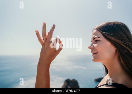 Frau Geste Ozean Junge Frau zeigt ein Zeichen in Ordnung, während sie lächelt und das Meer von einer Klippe an einem sonnigen Tag aus überblickt Stockfoto