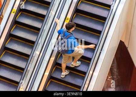 Hochwinkel-Rückansicht eines lässig gekleideten jungen Mannes mit Rucksack, der in einem modernen öffentlichen Gebäude eine Rolltreppe hinaufgeht. Stockfoto