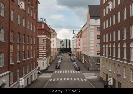 HELSINKI, FINNLAND - 11. AUGUST 2025: Lange, von Bäumen gesäumte Wohnstraße mit rot-weißen Wohngebäuden, die sich zu einem Fluchtpunkt zurückziehen. Stockfoto
