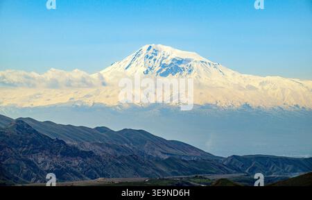 Der schneebedeckte Berg erhebt sich über fernen Hügeln unter klarem blauen Himmel. Der Berggipfel glänzt weiß, im Kontrast zu den felsigen Unterhängen. Wolkenschichten Stockfoto