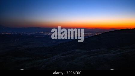 Die Lichter der Stadt leuchten in der Abenddämmerung von einem Hügel aus. Der orangefarbene Sonnenuntergang verblasst in den tiefblauen Himmel. Sanfte Hügel umrahmen eine ferne Stadtlandschaft. Keine Tiere oder Pflanzen Stockfoto