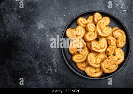 Klassische französische palmier-Kekse aus Blätterteig in schwarzer Schüssel dunkler Hintergrund süß gebackenes Dessert-Konzept. Stockfoto