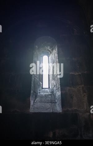 Dunkle Steinmauer mit schmalen Bogenfenstern. Licht leuchtet durch die enge Öffnung. Antike Architektur mit strukturiertem Mauerwerk. Atmosphärischer Blick auf Aban Stockfoto