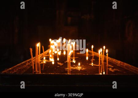 Viele Kerzen leuchten in der Dunkelheit warm. Die Flammen flackern sanft auf der Holzoberfläche. Kerzenhalter stehen aufrecht mit sanftem Licht. Warmes Ambiente geschaffen Stockfoto