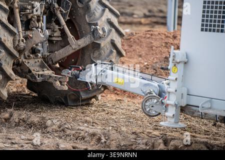 Die Heckkupplung des Traktors ist mit einem weißen Industrieanhänger mit Stützrad, rauem Boden und Maschinenkupplung für die Baustellenlogistik verbunden Stockfoto
