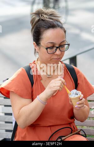 Frau im Alter von 45 Jahren hält einen Löffel und Eis, während sie sich auf einer Stadtbank ausruht Stockfoto