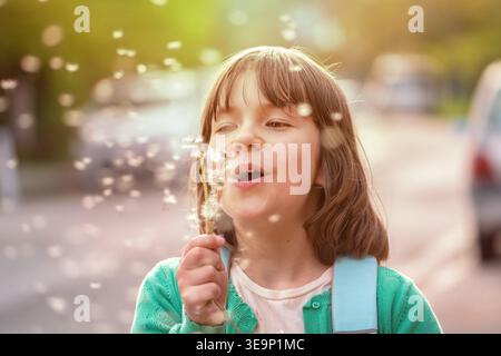 8 Jahre altes Kind, das Löwenzahnsamen im Sonnenlicht auf Stadtstraßenhintergrund freisetzt. Stockfoto