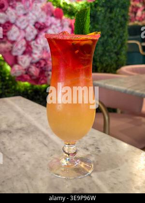 Vibrierender Cocktail in Rot- und Orangetönen, garniert mit Minze, serviert in einem hohen Glas auf einem Marmortisch mit floralem Hintergrund. Stockfoto