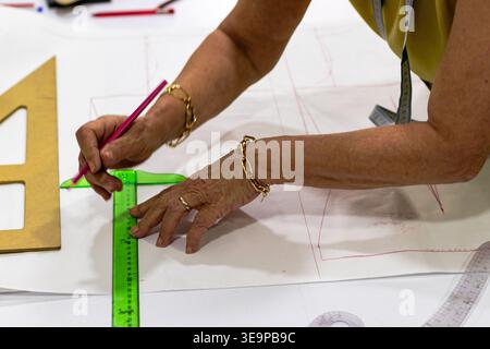 Die Hände eines Schneiders zeichnen gekonnt Linien auf Musterpapier mit einem rosafarbenen Bleistift und einem grünen Lineal, mit einem goldenen Zeichnungsdreieck in der Nähe. Stockfoto