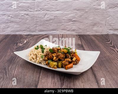Ein gesundes, hausgemachtes Hühnchen-Reis-Gericht mit Gemüse, serviert auf einem weißen Teller auf einem rustikalen Holztisch Stockfoto