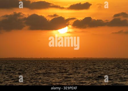 Ein faszinierender Blick auf den Sonnenuntergang am Horizont, der goldene Farbtöne über dem Meer ausstrahlt. Der leuchtende orange Himmel ist teilweise von weichen Wolken bedeckt Stockfoto