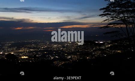 Panoramablick auf Bucaramanga, Kolumbien, beleuchtet bei Nacht unter einem tiefblauen Dämmerhimmel, eingerahmt von Baumsilhouetten und verblassenden Farben des Sonnenuntergangs Stockfoto
