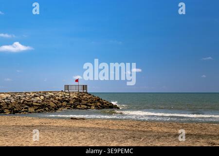 Ein ruhiger Blick auf einen sonnigen Strand in Salgar, Kolumbien, mit einem felsigen Wellenbrecher und einer roten Flagge, die Vorsicht signalisiert. Stockfoto