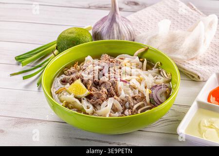 Eine leuchtend grüne Schüssel mit traditioneller vietnamesischer Rindfleischnudelsuppe Pho bo, mit Reisnudeln, Sojasprossen und Zwiebeln. Serviert mit frischer Limette, Chili, an Stockfoto