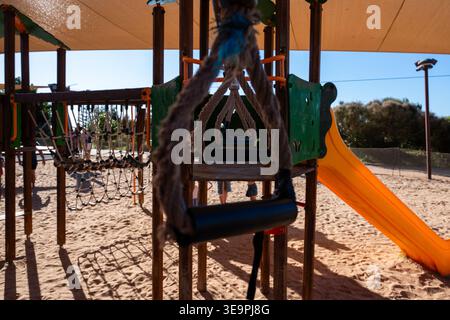 Der sonnendurchflutete Spielplatz verfügt über eine Schaukel und Rutsche. Sanddünen umrahmen die Szene unter einem Baldachin. Stockfoto