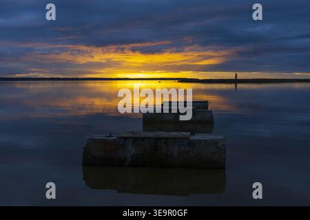 Goldener Sonnenuntergang reflektiert auf stillen Wasser mit Betonkonstruktionen im Vordergrund. Minimale atmosphärische Küstenszene. Stockfoto