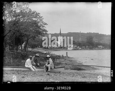 C.J. Merfield (F.R.A.S), F.K. McClean (F.R.A.S) und Captain G.H. Lacy sind auf diesem Foto aus dem Jahr 1908 zu sehen, das an einem Strand in Papeete, Tahiti, aufgenommen wurde. Zwei Männer sitzen auf einem weißen Kanu, während ein Drittel leicht erhöht hinter ihnen sitzt. Eine Kirche und Fahnenmast sind im Hintergrund zu sehen, mit Bäumen um die Gegend und einem Hügel in der Ferne. Stockfoto