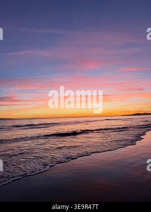 Ein farbenfroher Sonnenuntergang über sanften Meereswellen, die sich auf einen Sandstrand stürzen. Stockfoto