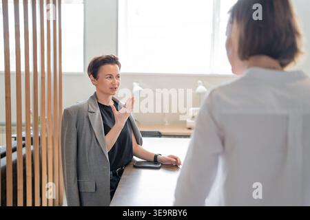 Zwei Frauen diskutieren im modernen offenen Büro mit Stehtisch mit Holzlamellen. Diskutieren, Stehen, Trennen, Büro, Teamarbeit, Frauen. Stockfoto