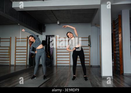 Zwei Frauen führen seitliche Dehnübungen in einem Fitnessstudio durch. Sie folgen gemeinsam einer Fitness-Routine. Der Raum ist hell und für Workouts konzipiert. Stockfoto