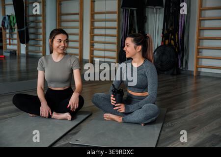 Zwei Frauen sitzen auf Yogamatten in einem Fitnessstudio. Sie lächeln und reden nach dem Training. Der Raum verfügt über Fitnessgeräte und Spiegel. Stockfoto