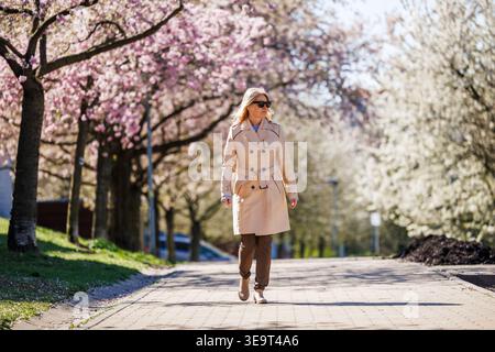 Frau mit Sonnenbrille im Trenchcoat spaziert im Frühling auf dem Gehweg unter der blühenden Sakura-Gasse in der Stadt. Urbaner Lebensstil Stockfoto