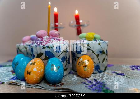 Lebendige, Bemalte Eier Um Dekorierte Kuchen Und Beleuchtete Kerzen Auf Gemustertem Tuch, Strukturierte Zuckerglasur Und Blumenmotive Sorgen Für Einen Gemütlichen Frühling Stockfoto