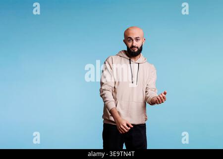 Junger arabischer Mann in guter Laune tanzt und in die Kamera schaut. Entspannter kahlköpfiger Bärtiger in lässigem Hoodie, der Hände und Körperbewegungen macht, Rhythmus spürt und ein Studio-Porträt feiert Stockfoto