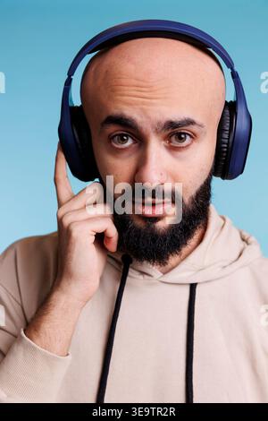 Junger arabischer Mann, der Musik im Porträt kabelloser Kopfhörer hört. Glatzbärtige Person, die Musik-Playlist in Ohrhörern genießt, während sie mit neutralem Ausdruck in die Kamera schaut Stockfoto