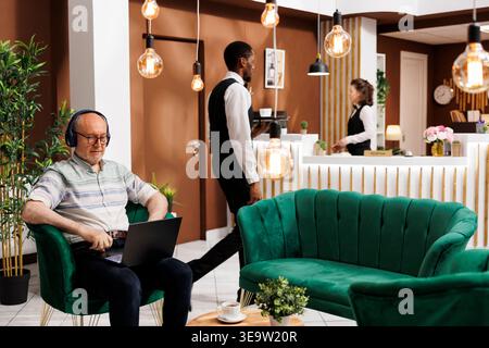 Ein älterer Rentner trägt Kopfhörer und sitzt mit seinem Computer in der Hotellobby. Ein älterer männlicher Reisender mit schnurlosem Headset wartet im Loungebereich und nutzt einen Laptop, um im Internet zu surfen. Stockfoto