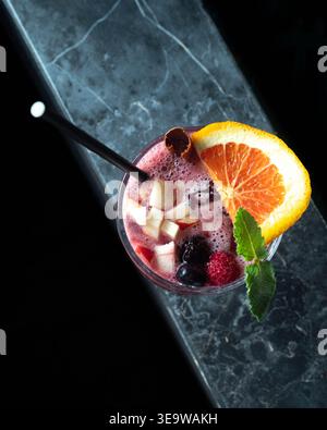 Frisches Beerengetränk garniert mit Orange und Minze. Schwarzer Strohhalm liegt in einem lebendigen, schaumigen Getränk. Die Marmoroberfläche sorgt für einen eleganten Kontrast zum dunklen Hintergrund Stockfoto