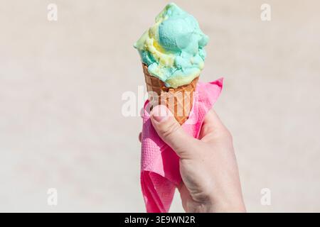 Die Hand hält einen Waffelkegel gefüllt mit farbigem Eis in einer knalligen rosafarbenen Serviette. Der sanfte, neutrale Hintergrund unterstreicht den erfrischenden Sommer Stockfoto