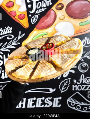 Goldene, gegrillte Käsescheiben sitzen auf einem Holzbrett. In zwei kleinen Schüsseln können eingetauchte Saucen aufbewahrt werden. Der Hintergrund der Kreidetafel mit Käse sorgt für zusätzlichen Spaß. Das Essen sieht toll aus Stockfoto