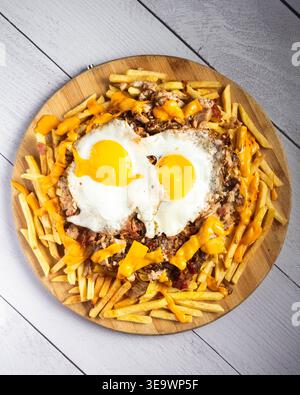 Herzförmige Eier sitzen auf knusprigen Pommes Frites und geschmolzenem Käse. Goldene Pommes Frites umgeben das Gericht auf einer rustikalen Holzplatte. Helles, sonniges Eigelb Stockfoto