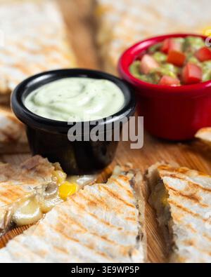 Goldene Quesadillas auf Holzbrett aufgespalten. Cremige Soße und frische Salsa in kleinen Schüsseln. Strukturierte Grillkanten stehen im Kontrast zu einer weichen, käsigen Füllung. Stockfoto