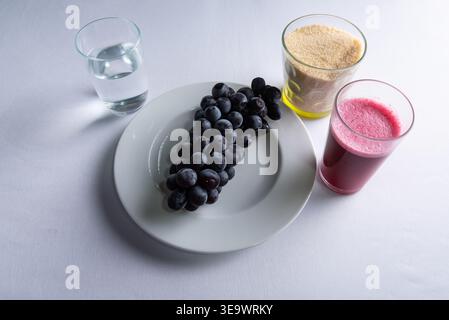 Ein Haufen dunkler Trauben auf weißem Teller mit Wasserglas, Zuckerglas und dunkelrosa Saftglas. High Key Product Shot liefert saubere, helle Lebensmittel und Be Stockfoto