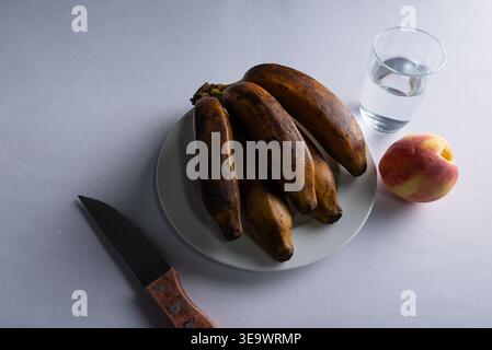 Still Life verfügt über überreife Bananen auf weißem Teller, Pfirsich- und Wasserglas auf weißem Teller. Die sanfte Studiobeleuchtung erzeugt Schatten, die Texturen freigeben. Diät, Heilung Stockfoto