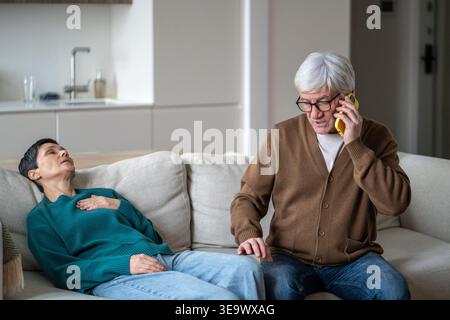 Eine ältere Frau lag auf der Couch im Zimmer mit Schmerzen in der Brust, während der Mann am Telefon mit dem Arzt sprach. Stockfoto