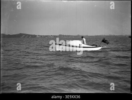 Irene May, fotografiert am 23. März 1912, wird auf dem Wasser mit einem Mann an Bord gezeigt. Die neuseeländische Flagge fliegt vom Heck aus, mit einem Küstenabschnitt im Hintergrund. Stockfoto
