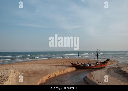 Ein kleines, verwittertes rotes Fischerboot sitzt in einem flachen Gezeitenkanal an einem Sandstrand, an dem die sanften Wellen des Ozeans auf den Horizont treffen. Stockfoto