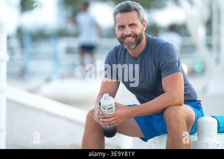 Sport-Fitness-Mann, der draußen auf einer Bank ruht. Reifes Fitnesssportmodell, das nach dem Training auf einer Bank sitzt. Hispanischer Sportmann macht danach eine Pause Stockfoto