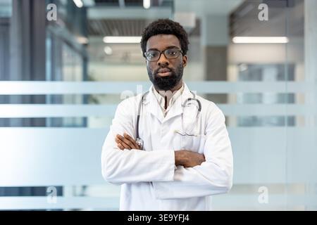 Selbstbewusster junger männlicher Arzt, der mit überkreuzten Armen in einem Krankenhaus oder einer Klinik steht, einen weißen Labormantel und ein Stethoskop trägt, was die Professionalität des Gesundheitswesens und die medizinische Expertise repräsentiert Stockfoto