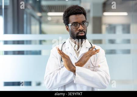Junge Ärztin, die Verbot und Ablehnung ausdrückt, mit gekreuzten Armen einen Stopp oder eine Ablehnung anzeigt, einen weißen Labormantel und Stethoskop in einer modernen Klinik trägt Stockfoto