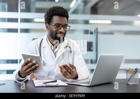 afroamerikaner Arzt mit Labormantel und Stethoskop. Lächeln, während Sie ein digitales Tablet und einen Laptop am Schreibtisch verwenden. Bereitstellung virtueller medizinischer Beratung und Nutzung moderner Gesundheitstechnologie Stockfoto