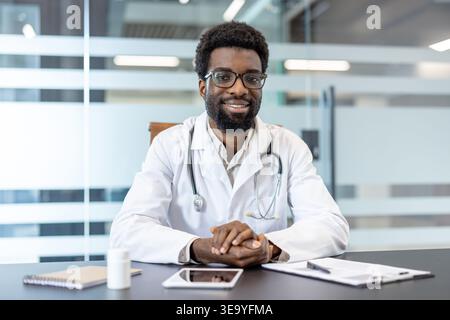 Freundliches medizinisches Fachpersonal lächelt und blickt in die Kamera und bietet zuverlässige medizinische Beratung, während Sie an einem Schreibtisch in einem modernen Klinikbüro sitzen Stockfoto