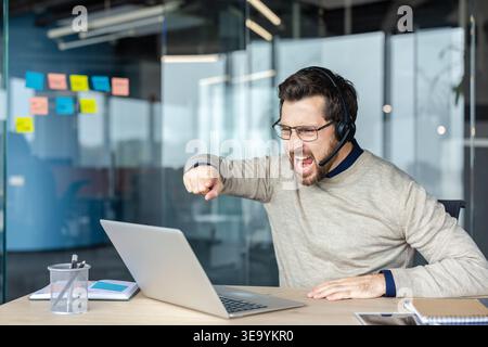 Gestresster männlicher Mitarbeiter, der ein Headset und eine Brille trägt und Wut und Frustration ausdrückt, einen Laptop anschreit und Burnout erlebt, während er in einer modernen Büroumgebung arbeitet Stockfoto